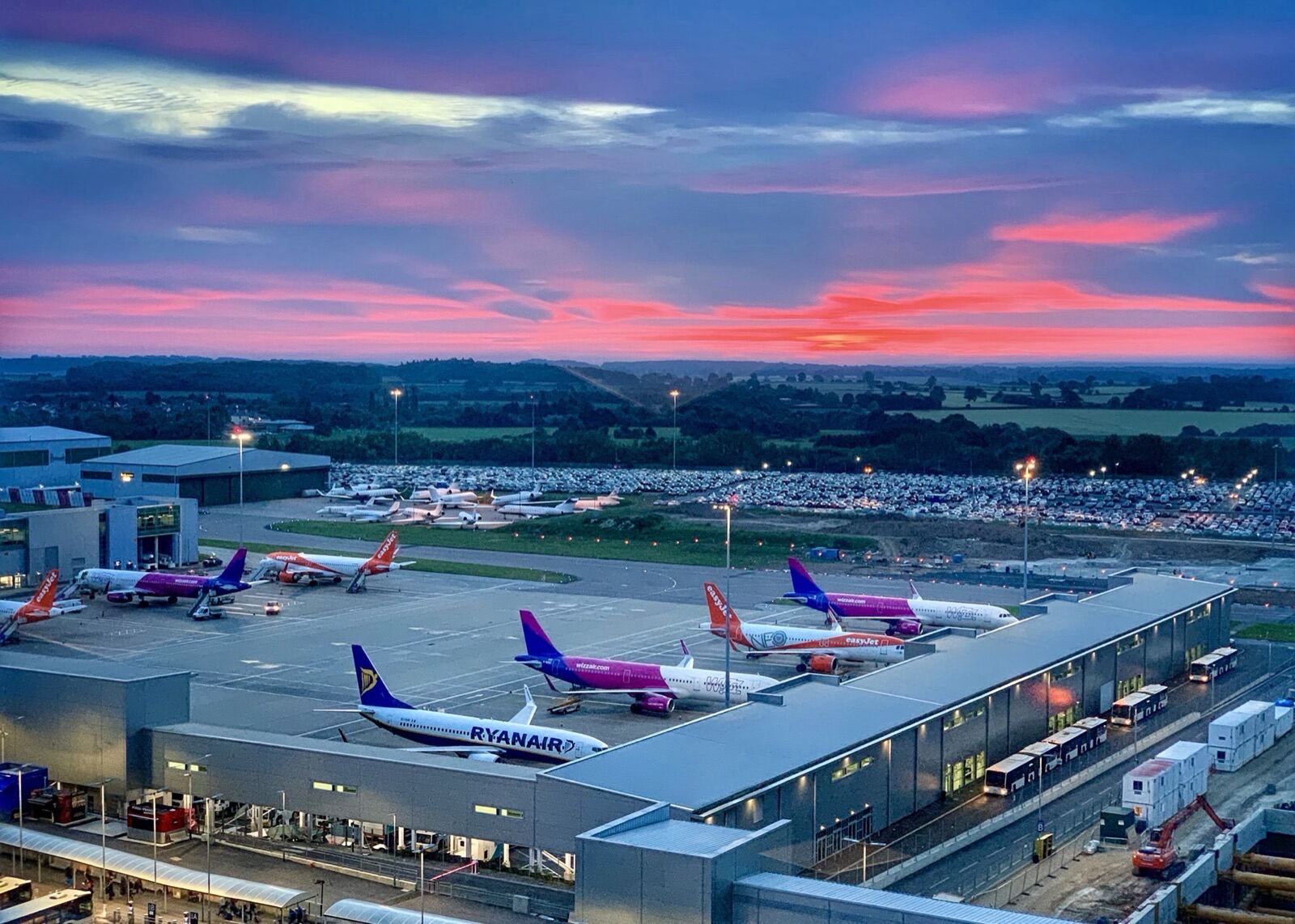 An airport apron at dusk, aircraft at their stands under a pink sky
