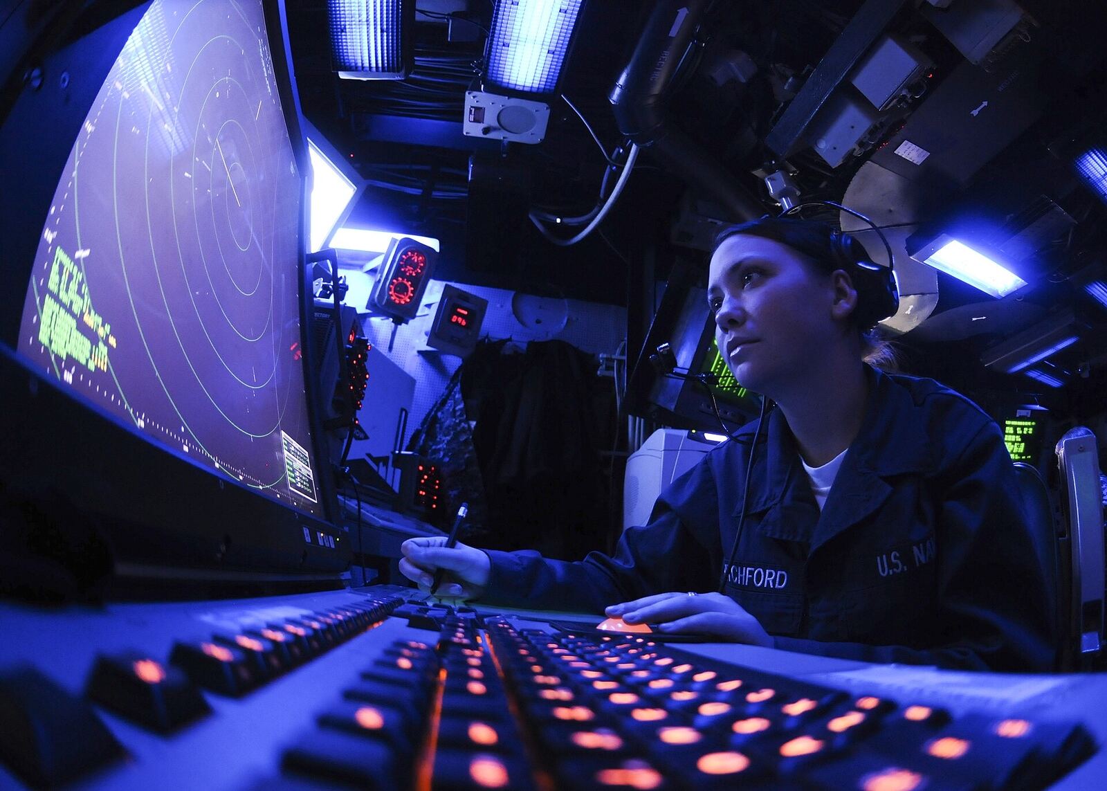 A radar operator at a console lit in blue, a radar screen to her left