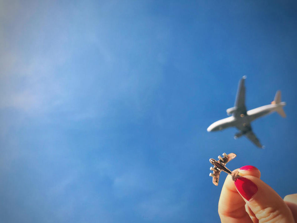 A hand holding a small aircraft pin up against a blue sky, an airliner passing overhead